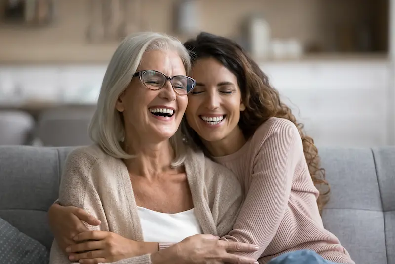 A happy dental patient laughs while being hugged by her daughter on a couch.